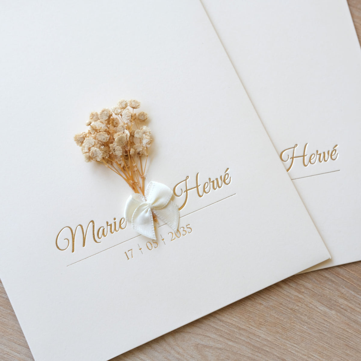 Détail de la pochette crème d'un faire-part de mariage avec prénoms en dorure, mini-bouquet de fleurs séchées et nœud en satin blanc.